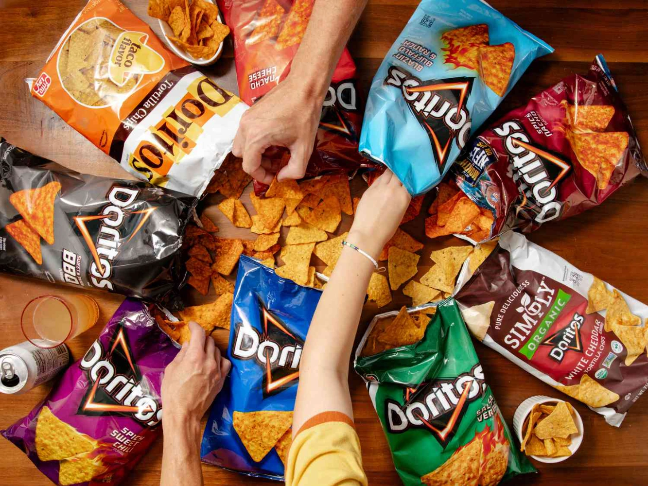 Multiple Doritos flavor bags spread on a table with people reaching for chips